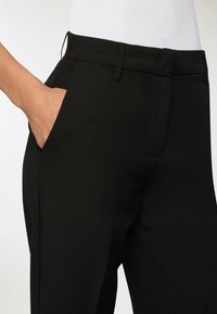 Czarne spodnie z długimi nogawkami o gładkiej strukturze, prostym kroju i bocznymi kieszeniami. Szczegóły to szlufki na pasek oraz czysty pas.
