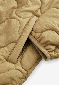 Manche de veste en tissu beige matelassé avec couture apparente et un bord de manche simple, mettant en valeur la texture et les détails des coutures.