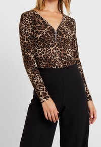 Bodysuit s leopardím potiskem, dlouhými rukávy, ve výstřihu do V a se zipem, se zaobleným akcentem. V kombinaci s černými kalhotami s vysokým pasem.