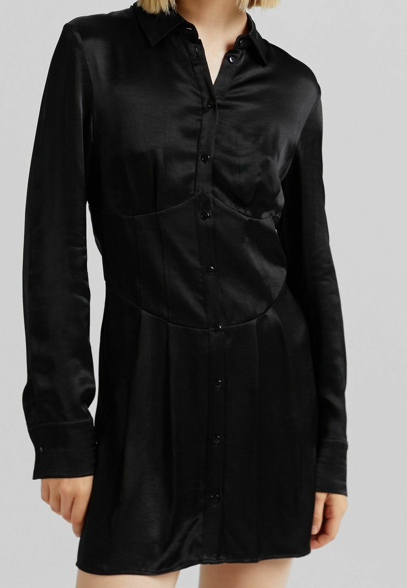 Femme portant une robe-chemise noire en satin à manches longues, boutonnée devant et col, debout devant un fond uni gris clair.
