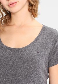 Šedé tričko s krátkým rukávem a kulatým výstřihem, vyrobené z měkké tkaniny. Obsahuje malé logo na hrudi v červené, bílé a modré barvě.