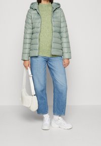 Polstret, lysegrøn jakke med hætte, struktureret grøn striktrøje, blå jeans med lige ben, hvid strikket taske og hvide sportssko.