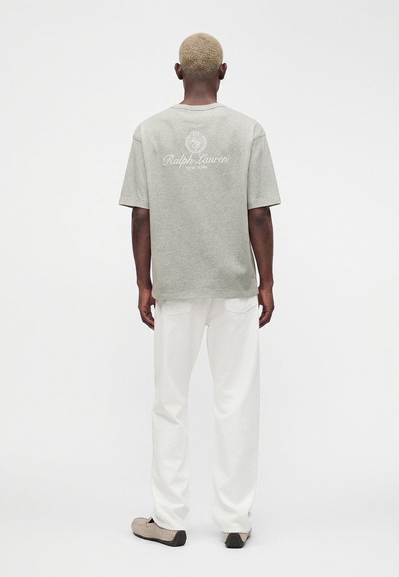 Personne aux cheveux blonds courts, vue de dos, portant un t-shirt gris Ralph Lauren, un pantalon blanc et des chaussures beiges sur un fond uni.