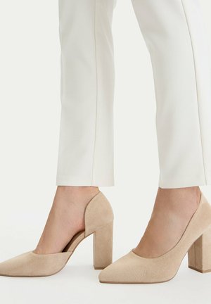Beige Wildleder-Pumps mit Blockabsatz, getragen zu weißen schmal geschnittenen Hosen, mit seitlichem Ausschnittdetail und spitzen Zehen.