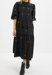 Abito nero con maniche a sbuffo corte, fiori blu ricamati, balze arruffate e chiusura a bottoni sul davanti. Abbinato a stivali neri alla caviglia.