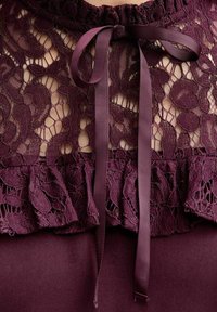 Blusa burdeos con encaje superpuesto que presenta un dobladillo con volantes y un lazo en el cuello. El encaje tiene un patrón floral con un acabado texturizado.