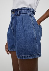 Denim-korkeastuvut, joissa on poimutuksia, sivutaskut ja nappikiinnitys, yhdistettynä sisätakkiin valkoiseen paitaan.