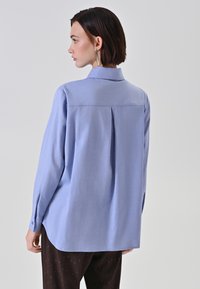 Camicia a maniche lunghe azzurro chiaro con colletto, dotata di una piega sul retro, polsini con bottone e orlo arrotondato. Tessuto liscio con una texture sottile.