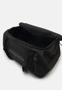 Black duffel bag with a zippered top, featuring a spacious interior, adjustable straps, and a prominent gray "patagonia" logo on the side.