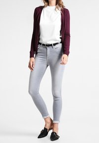 Bordowy kardigan, biała koszulka, jasnoszare obcisłe dżinsy i czarne wsuwane buty z klamrami. Gładkie tkaniny i dopasowany fason.