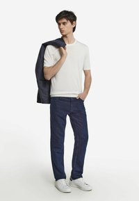 Vit kortärmad skjorta, marinblå jacka slängd över axeln, mörkblå jeans och vita sneakers. Enkel, figurnära design med minimal detalj.