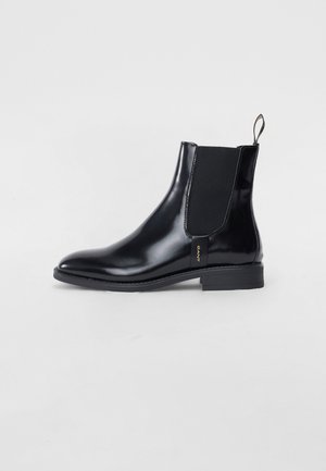 Botín Chelsea de cuero negro con paneles laterales elásticos, punta redondeada y un tacón bajo. Presenta una lengüeta decorativa y una sutil marca.