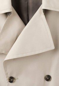 Beżowa tkanina płaszcza trencz z dużym kołnierzem, dwurzędowym fasonem, dwoma ciemnymi guzikami i gładką satynową podszewką.