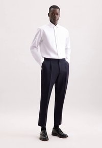 Wit overhemd met knopen en een brede kraag, gecombineerd met marineblauwe, op maat gemaakte pantalon. Model draagt zwarte leren schoenen. Glad, nauwsluitend materiaal.