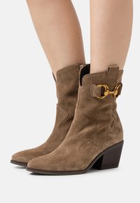 Botas de tobillo de ante marrón con punta afilada, diseño esculpido y un hebilla de acento dorado en el lateral. Tacón mediano apilado.