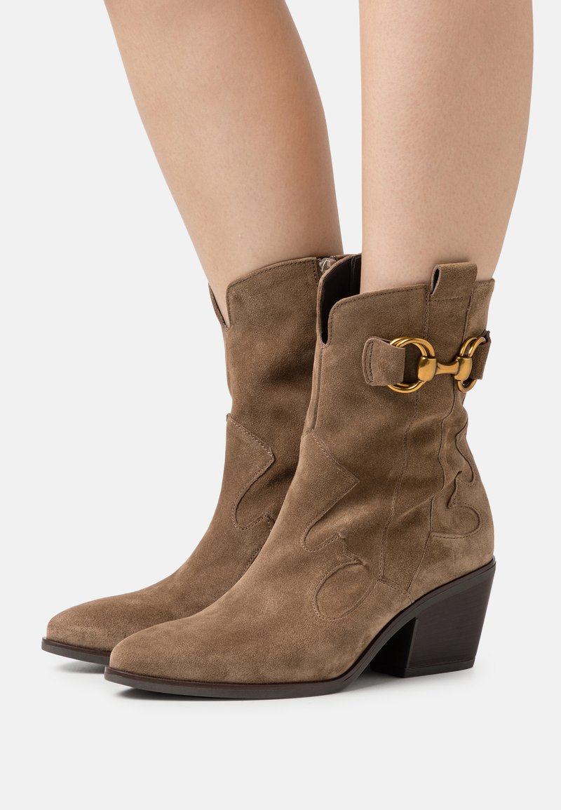 Botas de tobillo de ante marrón con punta afilada, diseño esculpido y un hebilla de acento dorado en el lateral. Tacón mediano apilado.