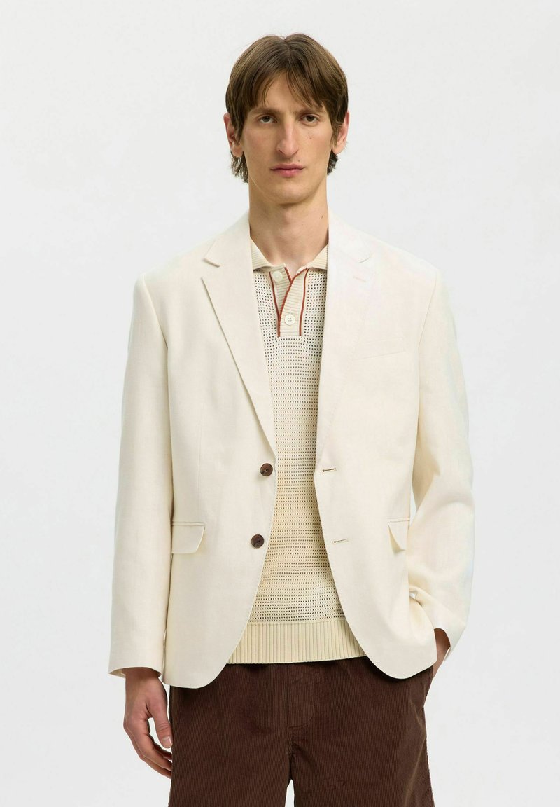 Homme portant un blazer crème par-dessus un polo en maille beige et un pantalon en velours côtelé marron, debout devant un fond blanc uni.