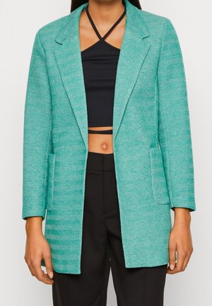 Femme portant un blazer en chevrons couleur sarcelle par-dessus un crop top noir à bretelles halter et un pantalon noir taille haute, se tenant devant un fond uni.