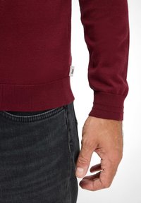 Burgunderfarbener Strickpullover mit gerippten Bündchen, kombiniert mit dunklen Jeans; verfügt über ein kleines Stoffetikett am Saum.