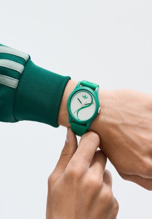 Grüne Silikonarmbanduhr mit einem weißen Zifferblatt, das ein geschwungenes Design und minimalistische florale Akzente aufweist. Verstellbares Armband mit Belüftungsöffnungen.