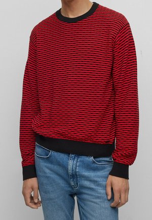 Homme portant un pull à rayures horizontales rouges et noires avec des poignets et un col noirs, associé à un jean en denim bleu.