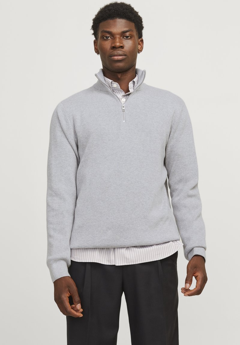 Man in een grijze quarter-zip trui over een gestreept overhemd, gecombineerd met zwarte broek, staand tegen een effen lichte achtergrond.