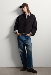 Veste noire courte avec des boutons, chemise grise à motif à carreaux aux poignets, jean bleu et baskets marron. Une casquette marron complète la tenue.
