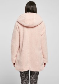 Abrigo de piel sintética rosa con una gran capucha, mangas largas y un corte relajado. Presenta una superficie texturizada y un dobladillo recto en la parte posterior.