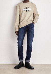 Sudadera beige con un logo negro en el frente, combinada con vaqueros azul oscuro arremangados en los puños y botines negros.