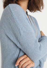 Ljusblå stickad tröja med avslappnad passform, med en V-ringning och en subtil silverdetalj längs kragen. Mjuk textur och ärmar i medellängd.