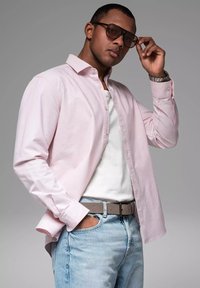 Lichtroze lange mouw shirt over een wit T-shirt, gecombineerd met lichtblauwe jeans en een grijze riem. Het model draagt donkere zonnebrillen.