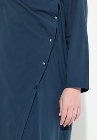 Il vestito blu navy presenta un design drappeggiato con una chiusura laterale con bottoni. Il tessuto morbido, le maniche lunghe e il dettaglio di cucitura visibile valorizzano il look.