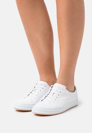 Jambes portant des baskets basses en cuir blanc avec des lacets blancs et des semelles en caoutchouc beige, debout sur une surface blanche.