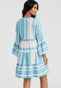 Vestido con patrones geométricos en azul y aqua sobre un fondo blanco, con mangas acampanadas y un diseño de falda en capas.