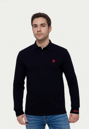 Joven vestido con una camiseta de manga larga negra con un logotipo rojo en el pecho y "USPA" en el puño, combinado con jeans azules, de pie frente a un fondo sencillo.