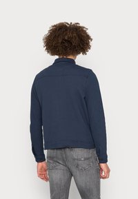 Tummansininen nyöri takki, jossa on kaulus, teksturoitu kangas ja nappiyksityiskohdat hihoissa, yhdistetty harmaisiin farkkuihin.