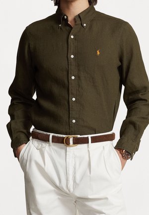 Homme portant une chemise boutonnée vert olive avec logo jaune, pantalon blanc plissé, ceinture marron avec boucle dorée et montre-bracelet, mains dans les poches.