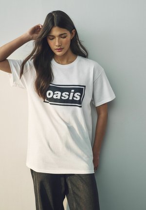 T-shirt en coton blanc avec le mot "oasis" imprimé en lettres noires grasses à l'intérieur d'un cadre rectangulaire. Coupe décontractée, manches courtes.