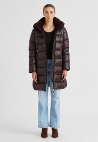 Frau in einem langen, dunkelbraunen Daunenmantel mit einem mit Fell gefütterten Kragen, schwarzem Oberteil, hellblauen Jeans und braunen Stiefeletten, die vor einem schlichten Hintergrund steht.