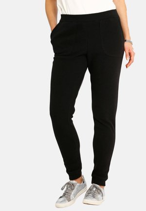 Frau trägt schwarze, eng anliegende Jogginghosen mit Vordertaschen, kombiniert mit silber-grauen Sneakers und einem weißen Oberteil.