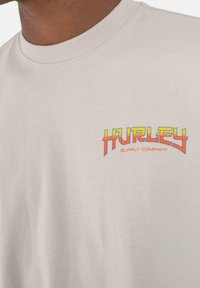 Camiseta de algodón color beige claro con un colorido logo de "Hurley Supply Company" en naranja, amarillo y rojo. Diseño de manga corta y cuello redondo.