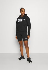 Czarna bluza z kapturem z białym logo Reebok, w połączeniu z czarnymi szortami z detalami logo. Model nosi czarne buty sportowe. Minimalistyczne tło.