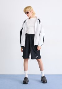 Veste coupe-vent blanche et noire, chemise blanche ajustée, shorts noirs et sabots noirs. Le modèle porte des lunettes de soleil teintées de vert, se tenant devant un fond bleu.