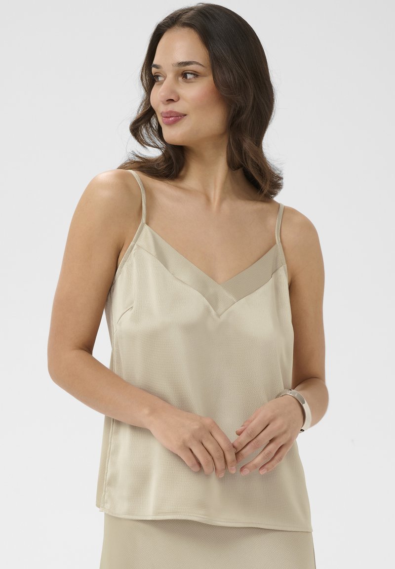 Femme aux cheveux ondulés châtain clair mi-longs, portant un haut beige sans manches à col en V et une jupe assortie, regardant sur le côté avec les mains doucement jointes.