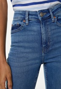 Jeans de mezclilla azul de tiro alto con un acabado desgastado, que presentan detalles metálicos de cobre, bolsillos frontales y cierre de cremallera.