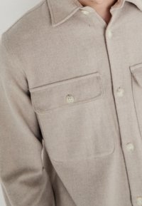 Beżowa koszula na guziki wykonana z mieszanki wełny, z ostro zakończonym kołnierzykiem, dwoma kieszeniami na piersi i jasnymi guzikami. Gładka faktura.