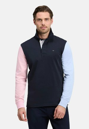 Homme portant un pull bleu marine avec une manche gauche rose et une manche droite bleu clair, se tenant devant un fond clair uni.