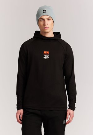 Jonge man die een lichtgrijze muts en een zwarte hoodie met een oranje "PROTEST" logo draagt, staand tegen een effen lichte achtergrond.