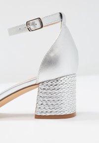 Silver sandal with a metallic finish, open back, ankle strap, and a textured block heel featuring a braided design.