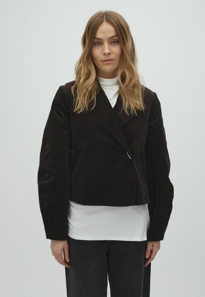 Femme aux cheveux blonds ondulés portant un blazer noir sur une chemise blanche, associée à un pantalon foncé, debout devant un fond clair uni.